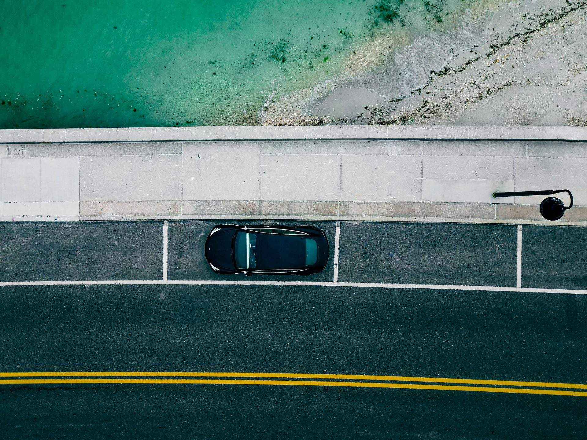 Vue du ciel : une voiture sombre garée le long d'une promenade, au bord d'une eau turquoise.
